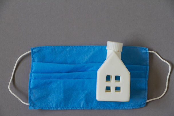 Fique em casa conceito, brinquedo figura da casa branca em azul máscara médica protetora, pano de fundo de quarentena
 - Foto, Imagem
