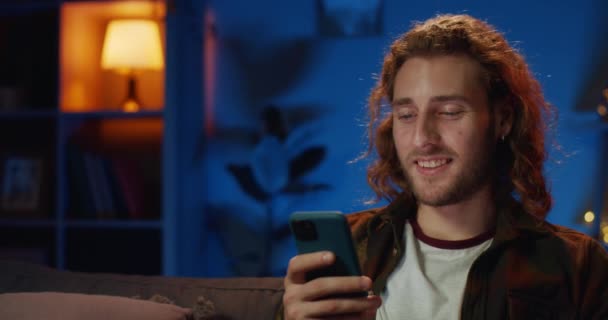 Close up view of millennial man with long hair using smartphone and laughing. Handsome bearded guy reading funny message and smiling while looking at phone screen at home. - Felvétel, videó