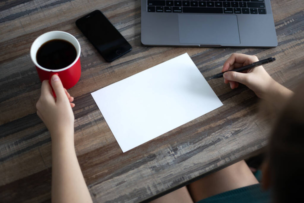 Handen schrijven op blanco papier met kopieerruimte met koffiebeker, telefoon, laptop op houten bureau tafel. Bovenaanzicht, close up - Foto, afbeelding