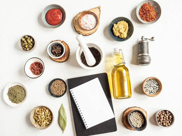 Kitchen notepad mockup for culinary text, various dry spices and sauces on light background. Flat lay of small bowls with dijon mustard, olive oil, ketchup, capers and spices. Top view - Photo, Image
