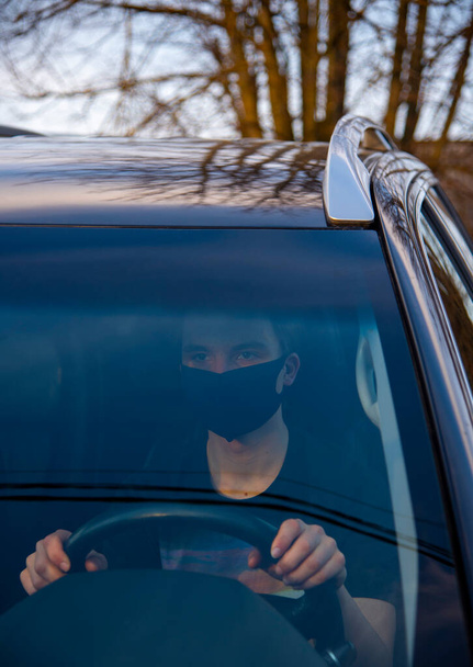 A young man in a medical mask is sitting at the wheel of a car. the photo was taken through glass. - Valokuva, kuva