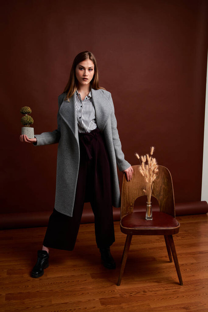 Mininalism style classic studio photo set of the young independent woman. Gorgeous young female in classic suit, black shoes and grey coat. Vintage chair. Woman's clothes. Fashionable clothes. - Фото, изображение