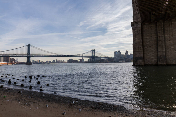 Brooklynský most z dálky, ze strany mostu, je vidět temná řeka a modrá obloha, za budovami města mostu - Fotografie, Obrázek