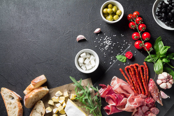 Tapas concept. Prosciutto, sausage chorizo, olives, cherry tomato, arugula, basil and brie cheese on a black slate board. Antipasti concept. Top view, copy space. - Photo, Image