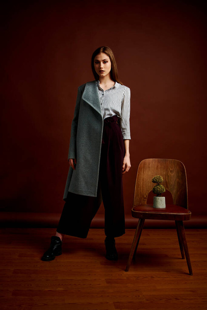 Mininalism style classic studio photo set of the young independent woman. Gorgeous young female in classic suit, black shoes and grey coat. Vintage chair. Woman's clothes. Fashionable clothes. - 写真・画像