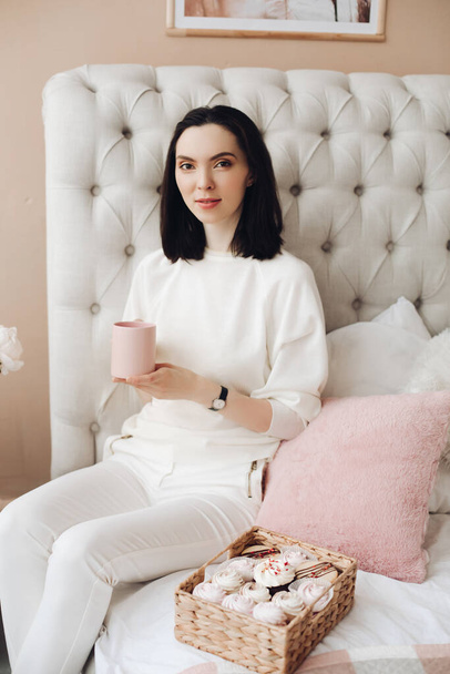 Bonita jovem se senta em uma cama grande com uma caixa de marshmallows
 - Foto, Imagem