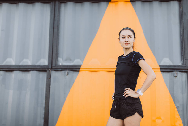 Girl dressed in a black T-shirt and shorts, warming up before running against the background of a yellow-gray wall. - Photo, Image