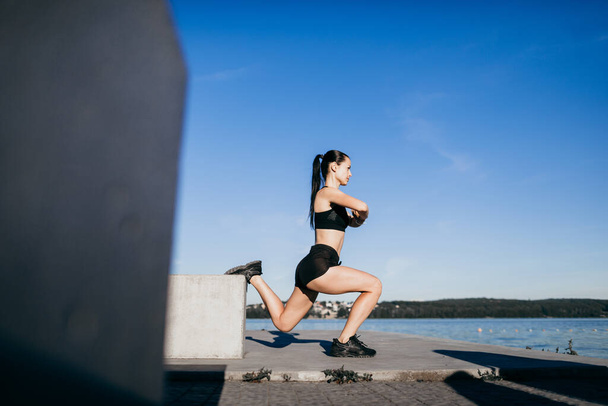 Siyah giyinmiş atletik kız egzersiz yapıyor, sabahları şehir plajında çömeliyor. - Fotoğraf, Görsel