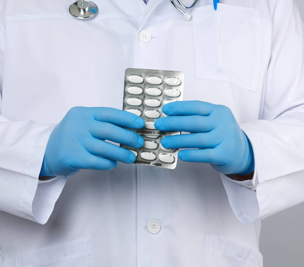 γιατρός θεραπευτής είναι ντυμένος με ένα λευκό ομοιόμορφη παλτό και μπλε αποστειρωμένα γάντια στέκεται και κρατώντας μια στοίβα από χάπια σε συσκευασίες κυψελών, λευκό φόντο - Φωτογραφία, εικόνα