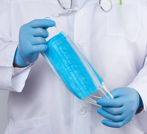lékař v bílém plášti, modré latexové sterilní rukavice drží v ruce textilní lékařské masky, ochranné doplňky proti virům a bakteriím, zblízka - Fotografie, Obrázek