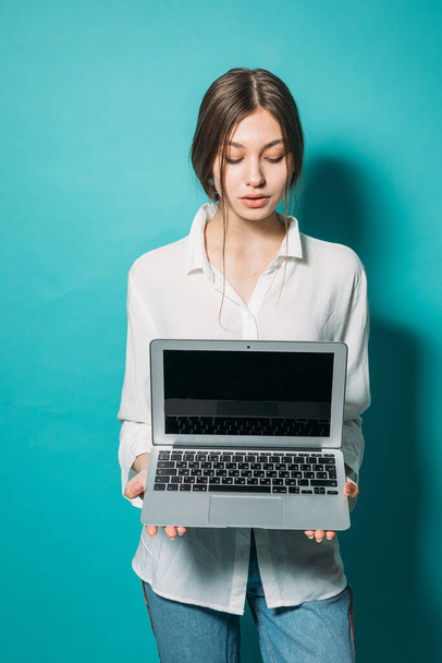 jovem menina em uma camisa branca e jeans com um laptop em um fundo azul no estúdio
 - Foto, Imagem