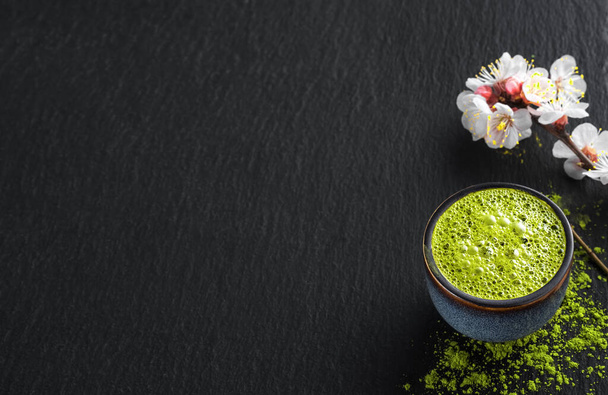 egy tál kék matcha zöld teával, egy virágzó cseresznye- és teapor mellett az asztalon. Teaidő, csend, fekete kő háttér másolási hely szöveg, lapos laikus - Fotó, kép