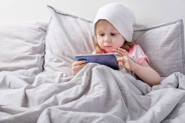 Baby in a white hat plays smartphone on the bed - Fotografie, Obrázek