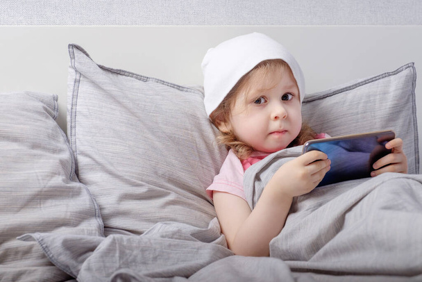 Baby in a white hat plays smartphone on the bed - Foto, Bild