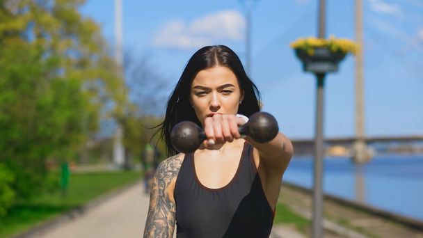 Fit brunette woman holds one old dumbbell during sunny day and looks at you - Foto, imagen