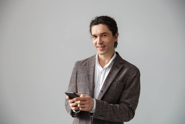 Image of pleased handsome man in jacket smiling and using cellphone isolated over white background - Zdjęcie, obraz