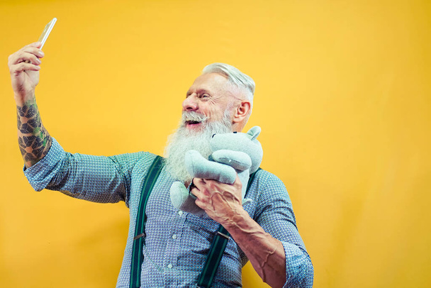 Portret starszego pana z misiaczkiem robiącym selfie. Człowiek i zabawka izolowane na żółtym tle. Koncepcja emeryta i technologii. Zdjęcie. - Zdjęcie, obraz