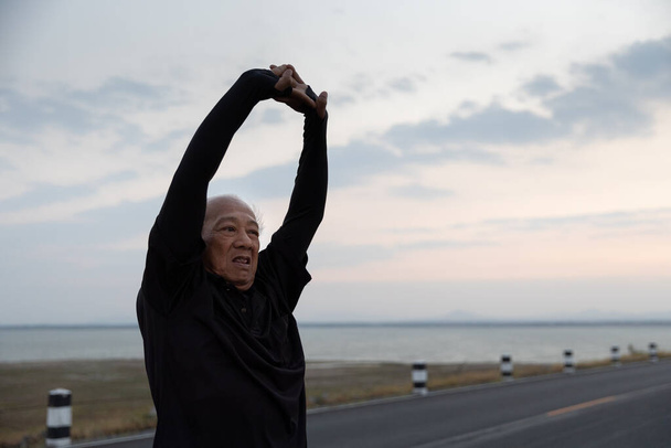 Ázsiai idősek gyakorlata. Idősebb férfi nyújtózkodó kar izmok és felkészülés futás gyakorlatok az úton a reggeli napsütés. Egészséges életmód és testmozgás, bemelegítés koncepció. Lassú mozgás - Fotó, kép