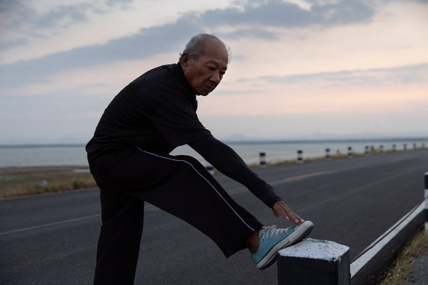 Asijské starší cvičení. Starší muž protahuje svaly nohou a připravuje se na cvičení na silnici v ranním slunci. Zdraví životní styl a cvičení, Zahřátí koncepce. Pomalý pohyb - Fotografie, Obrázek
