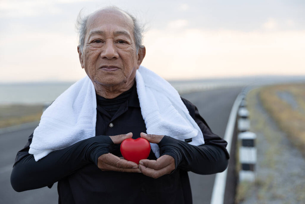 Asiático Hombre mayor en ropa de ejercicio sosteniendo el corazón rojo. Concepto Cuidado de la salud, Saludable con ejercicio y buen comienzo saludable, seguro, día mundial del corazón, seguro médico
 - Foto, Imagen