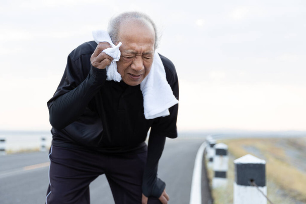 Cerca de ancianos asiáticos limpiando el sudor con una toalla después de hacer ejercicio corriendo por la carretera bajo el sol de la mañana. estilo de vida saludable y ejercicio Concepto
. - Foto, imagen