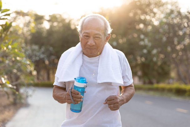 Front view Asian elderly exercise. Close up Senior man holding water bottle running on road in public park that has sunshine in the evening. health lifestyle and exercise Concept. - Photo, Image