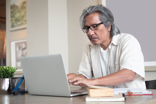 アジア系のシニア男性は、自宅の椅子に座っている間にノートパソコンを使用しています。高齢男性技術コミュニケーション。引退後の事業. - 写真・画像