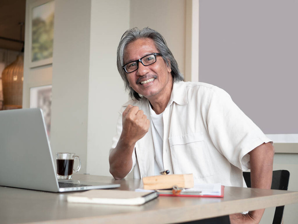 Ázsiai idősebb férfi laptoppal szemüveget visel, kamerába néz, és felemelt öklökkel diadalmaskodik, miközben otthon ül a széken. Üzleti sikerkoncepció - Fotó, kép