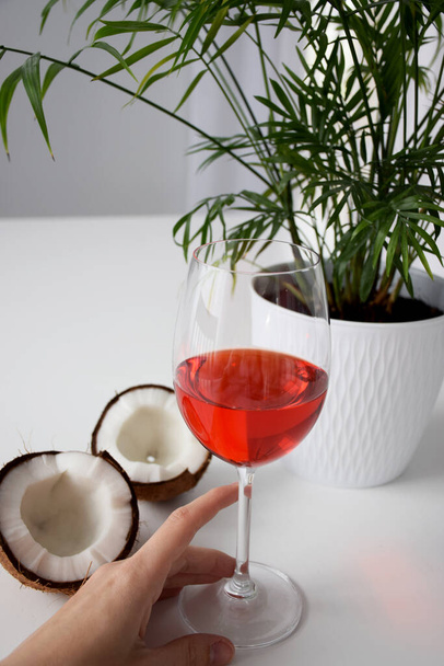 Cracked cocco vicino a casa pianta palma in una pentola bianca e un bicchiere di cocktail di vino con mano su un tavolo bianco durante il giorno
 - Foto, immagini