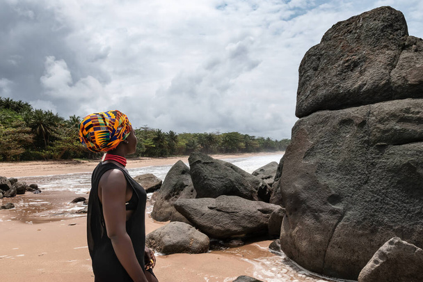 Donna africana in piedi su una bella spiaggia con grandi pietre scure e spiaggia pulita ai margini della giungla in Axim Ghana Africa occidentale - Foto, immagini