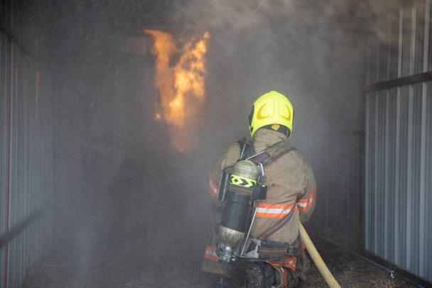 Achteraanzicht van de brandweerman draagt een brandbeveiligingspak dat de trainingsruimte binnenloopt en Spray water Fire op de brandweerkazerne. Training brandoefening Concept. - Foto, afbeelding