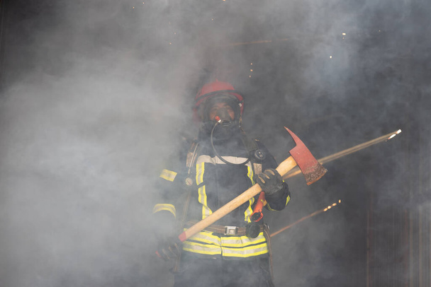 Азиатский пожарный держит Акса уверенно с дымом, огонь сильно горит на заднем плане. Концепция карьеры пожарного
. - Фото, изображение