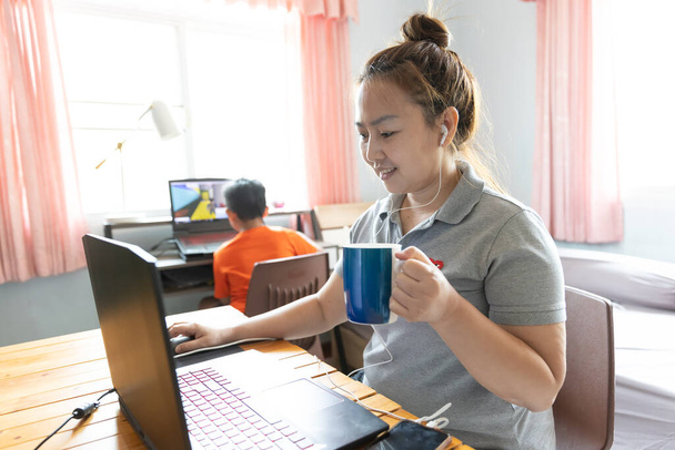 Mutlu Asyalı anne, sabahleyin oğlumla yatak odasında dizüstü bilgisayar kullanıyor ve kahve içiyor. Pencereden gelen güneş ışığı. Evden çalışmak. - Fotoğraf, Görsel