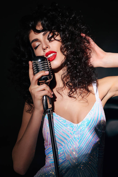 fashion studio photo of beautiful sexy woman with dark curly hair in elegant dress posing with microphone - Photo, Image