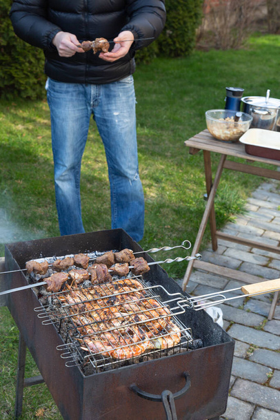 langoustines on grill and skewered meat are on barbecue with smoke on a green lawn outside. a man in a black down jacket and jeans strung pieces of meat on a metal skewer. - Foto, immagini