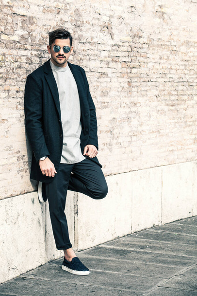 Knappe stijlvolle man leunend op een muur met een zonnebril en modern kapsel en baard. Buiten in de straat. Wit t-shirt en zwart jasje. - Foto, afbeelding