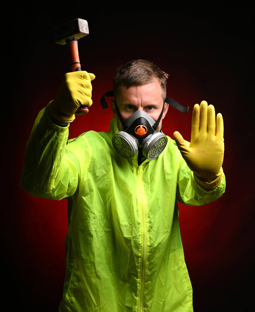 Man in respirator holding big hammer and showing STOP sign against dark red background - Foto, Imagen