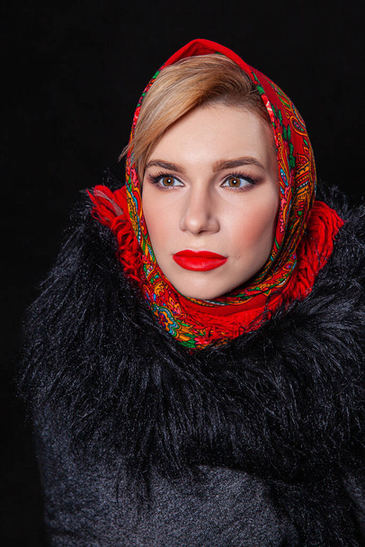 Uma linda loira com um olhar expressivo em um cachecol vermelho russo em sua cabeça e um casaco de pele preta em um fundo preto
 - Foto, Imagem