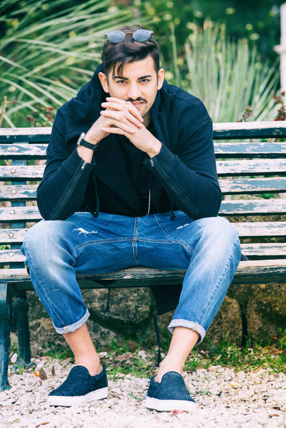 Handsome young man model sitting on the bench. A handsome young man is sitting on a bench watching and waiting. The guy wearing trendy clothes. Beside him the empty bench. - Foto, afbeelding