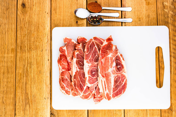 Chuletas de cerdo con condimentos en una tabla de cortar blanca sobre una mesa de madera, carne para barbacoa, vista superior, espacio para copiar, concepto de barbacoa
 - Foto, Imagen