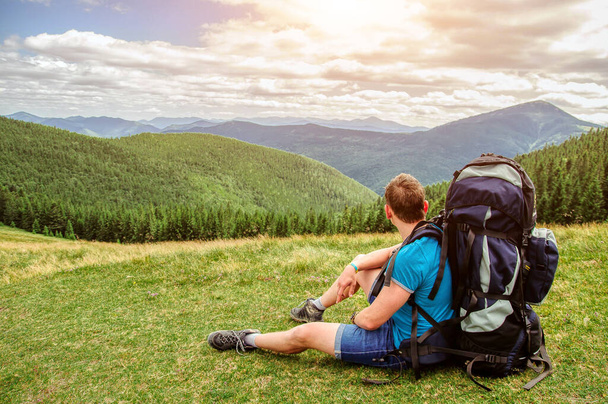 Joven turista con mochila sentado en la hierba y disfrutando de una hermosa vista de las montañas
. - Foto, imagen