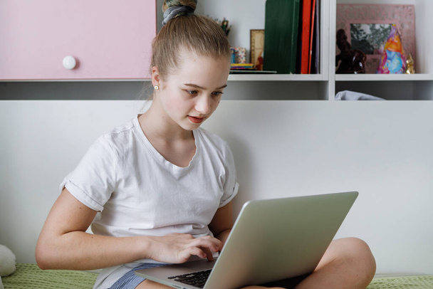 Teenager-Mädchen in weißem T-Shirt sitzt mit Laptop auf dem Bett. Schulbesuch zu Hause, soziale Distanz. Coronavirus unter Quarantäne. Selektiver Fokus. Hintergrund verschwimmen. - Foto, Bild