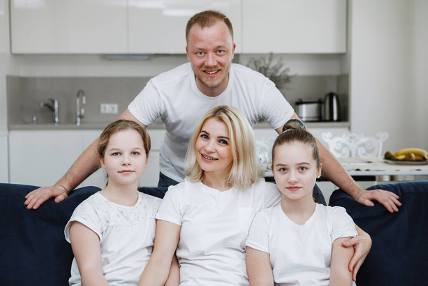 Négytagú fiatal család ül a kanapén. Apa, anya és két lánytestvér fehér pólóban mosolyognak és a kamerába néznek. A hajléktalanság. Szelektív fókusz. Homályos háttér. - Fotó, kép