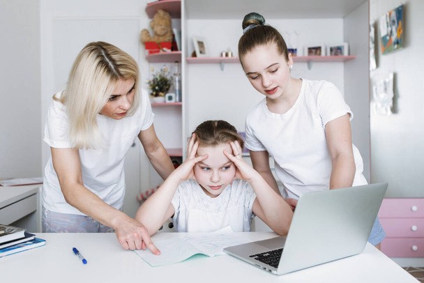 母親と妹は少女が宿題をするのを手伝う。十代の若者は動揺し、ノートパソコンでテーブルに座って、彼女の手の中に彼女の頭を握りしめました。隔離コロナウイルスだ。選択的フォーカス。ぼかしの背景. - 写真・画像