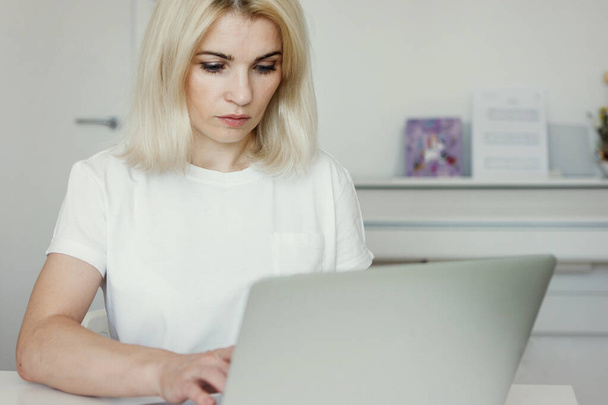 Blonde woman in a white T-shirt sits at a table and works with a laptop. Remote work. Quarantine coronavirus. Selective focus. Blur background. - Foto, immagini