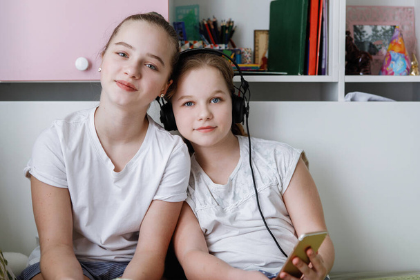 ヘッドフォン付きの白いTシャツを着た2人の10代の女の子がカメラを見て座っている。家にいる現代の若者. - 写真・画像