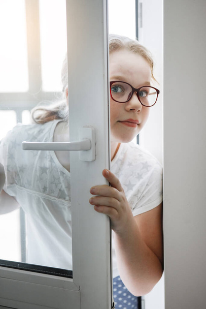 Adolescente avec des lunettes jette un coup d'oeil derrière une porte blanche ajar
. - Photo, image