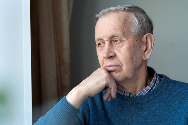 Porträt eines älteren Mannes. Ein nachdenklicher älterer Mann schaut aus dem Fenster und sitzt zu Hause. - Foto, Bild