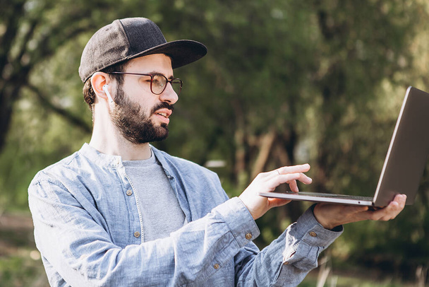 Attractive young man with dark hair in glasses works outdoors with a laptop on a sunny day. Bearded hipster guy wears casual clothes and works remotely at a quarantined company - Fotó, kép
