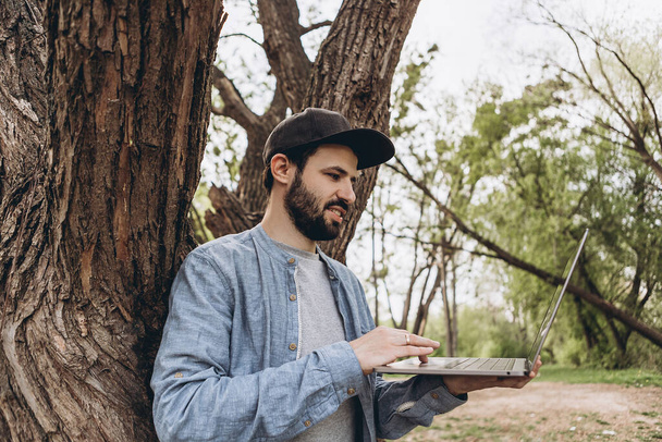 Attractive young man with dark hair in glasses works outdoors with a laptop on a sunny day. Bearded hipster guy wears casual clothes and works remotely at a quarantined company - Photo, image